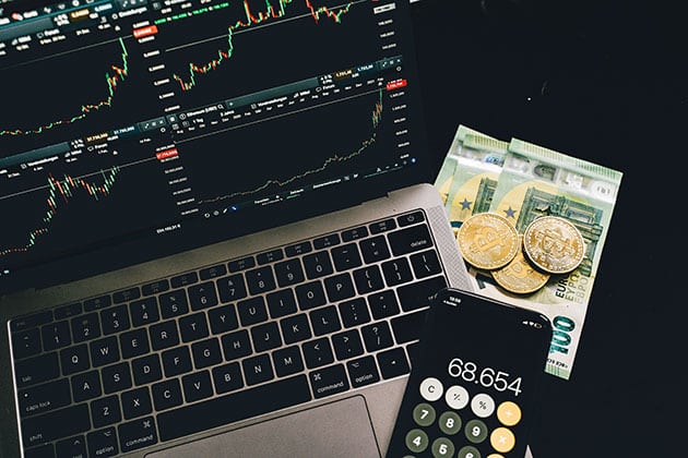 phone on a laptop showing crypto stocks, with cash and physical bitcoins beside it