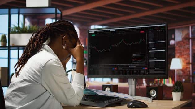 Woman at her desk with a monitor tracking crypto performance, holding her head.